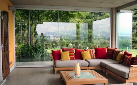 Come arredare una veranda aperta: un divano angolare grigio con cuscini rossi e gialli, un tavolino in legno e una vista sul giardino.