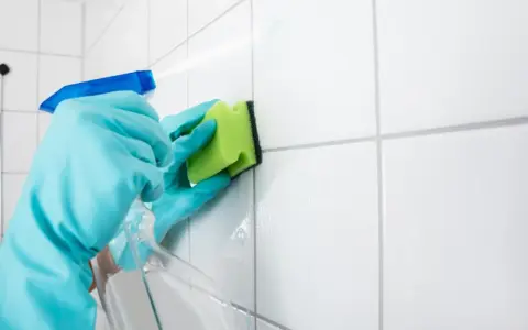 Manos con guantes azules rociando limpiador y frotando juntas de azulejos blancos con una esponja verde para blanquear juntas azulejos.