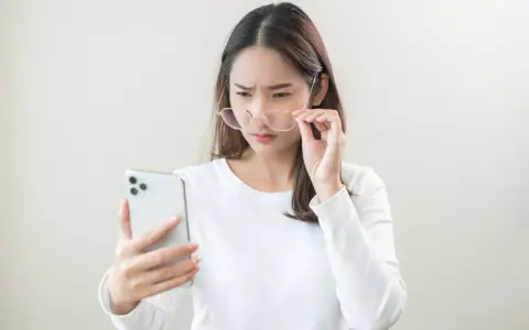 Kobieta z okularami próbuje odczytać coś na telefonie, marszcząc brwi. Może to być objaw astygmatyzmu krótkowzrocznego.