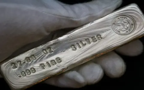 Mano guantata tiene un lingotto di **titolo argento** 999.9, con incisioni che indicano 27.00 oz e "FINE SILVER".