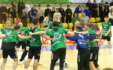 Krystian Woźniak i jego drużyna świętują zwycięstwo. Zawodnicy w zielonych strojach przytulają się na boisku, a na trybunach widać kibiców.