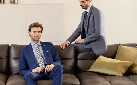 Due uomini eleganti in un lounge suit siedono su un divano di pelle marrone. Uno è seduto, l'altro è appoggiato.
