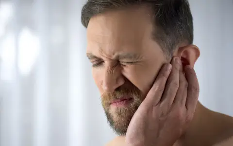 Mężczyzna z zarostem cierpi z powodu bólu chrząstki ucha przy dotyku, zaciskając oczy z bólu.