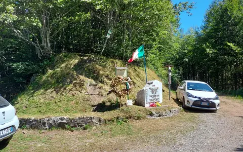 Monumento commemorativo con bandiera italiana e corona d'alloro al passo delle radici, circondato da alberi e con un'auto parcheggiata.