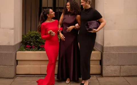Three women in elegant gowns, discussing what not to wear to a wedding. One in red, one in plum, and one in black.