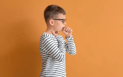 Chłopiec w okularach ma **szczekający kaszel u dziecka**. Dotyka gardła, czując dyskomfort.