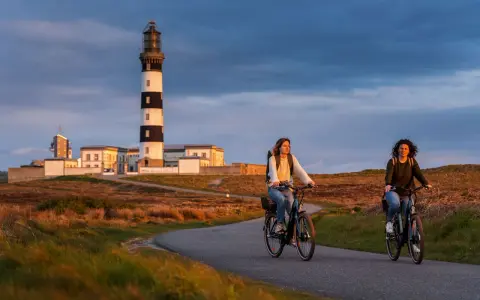 Deux femmes font du vélo sur une route sinueuse à Ouessant, avec un phare emblématique en arrière-plan. Une belle façon de visiter Ouessant à pied, ou plutôt à vélo !