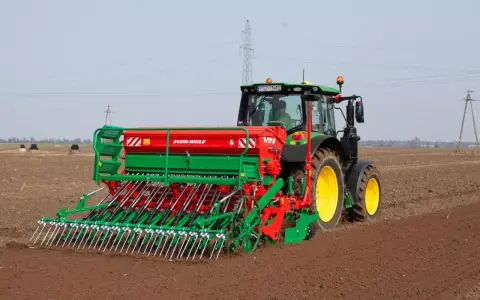 Zielony traktor z siewnikiem Agro-Masz pracuje na polu. Maszyna precyzyjnie wysiewa nasiona, optymalizując ile trawy na hektar zostanie zasiane.