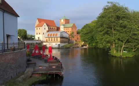 Zamek Darłowo z wieżami i zielonymi dachami, otoczony wodą i drzewami. Przy brzegu letnia restauracja z czerwonymi parasolami.