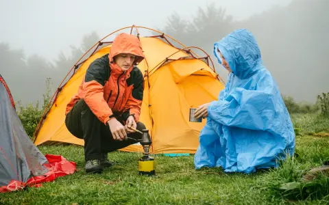 Zwei Camper in Regenponchos bereiten sich auf das Zelten bei Regen vor. Einer kocht auf einem Campingkocher, der andere hält eine Tasse.