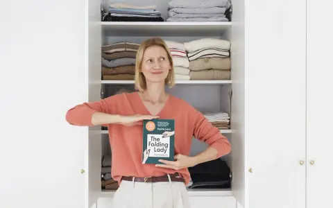 Kobieta z książką "The Folding Lady" pokazuje, jak składać koszulki, stojąc przed otwartą szafą pełną schludnie ułożonych ubrań.
