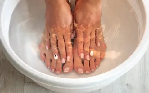 A relaxing home remedy foot soak in a white basin. Feet with french manicures and one toe nail decorated with a black and white design are submerged in clear water.