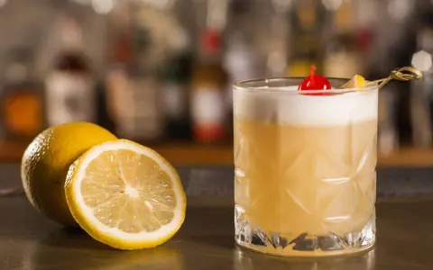 Orzeźwiający drink z amaretto, ozdobiony wisienką i plasterkiem cytryny, obok świeżych cytryn na barze.