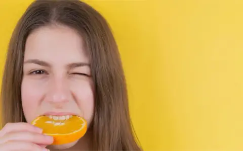 Junge Frau mit einem Stück Orange im Mund, die versucht, nach Weisheitszahn-OP wieder feste Nahrung zu essen.