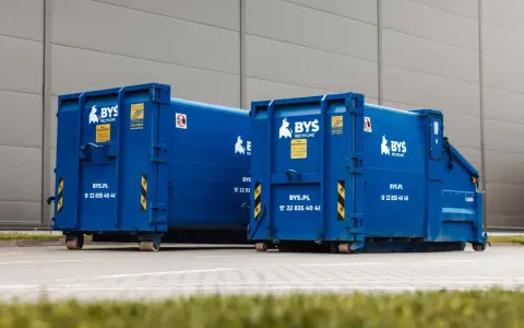 Dwa niebieskie kontenery BYS Recykling stoją przed budynkiem. Dowiedz się, ile kosztuje tona gruzu betonowego i jak go zamówić.