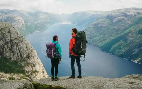 Två vandrare med stora ryggsäckar står på en klippa och blickar ut över en fjord i Norge. Perfekt för camping i Norge.