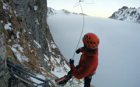 Wspinaczka górska w chmurach. Alpinista w pomarańczowym kasku i kurtce przygotowuje się do zjazdu na linie, otoczony zimowym krajobrazem i mgłą.