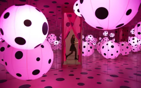 A woman walks through a mirrored room filled with pink polka-dotted spheres, a signature of Yayoi Kusama's immersive art.
