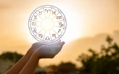 Dłonie trzymają koło zodiaku, gdzie widoczny jest symbol Wagi. Słońce przebija się przez niebo, tworząc magiczną aurę.