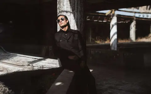 Hombre con gafas de sol y un **smart outfit** oscuro, apoyado en un pilar de hormigón en un entorno urbano abandonado.