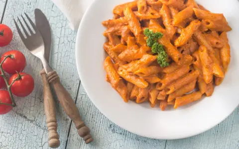 Piatto di penne alla vodka cremose, guarnite con prezzemolo, su un tavolo di legno azzurro. Accanto, pomodorini freschi e posate rustiche.