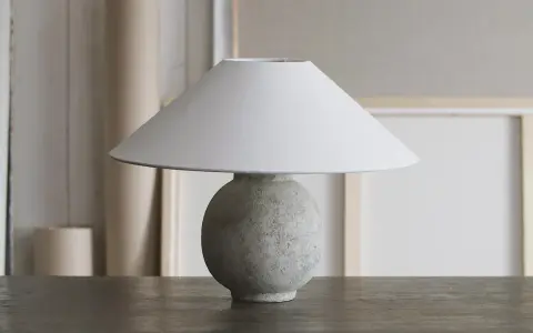 Bordslampa med rund, texturerad fot av lufttorkande lera, målad i grågröna toner. En vit lampskärm sitter ovanpå.