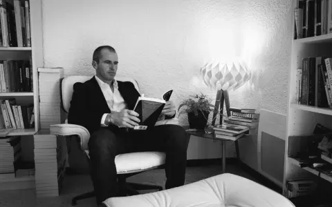 Un homme lit un livre dans un fauteuil, entouré de livres. La lumière d'une lampe éclaire la scène. ae-l af-l.