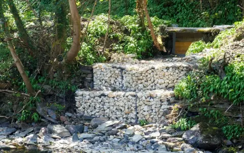 Estructura de muro de gavion con piedras blancas, rodeada de vegetación y rocas junto a un río.