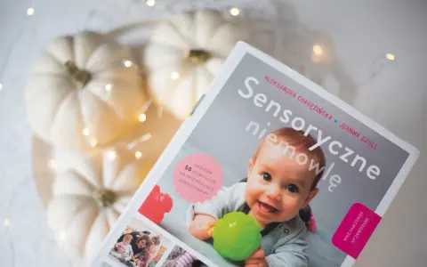 Okładka książki "Sensoryczne niemowlę" z uśmiechniętym dzieckiem, dyniami i lampkami.