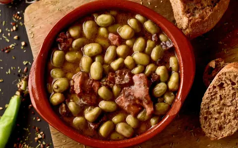 Un cuenco de barro con habas a la catalana, trozos de tocino y pan rústico al lado.