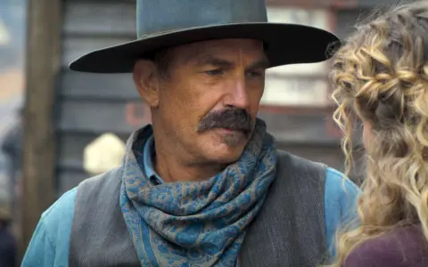 Kevin Costner w filmie z westernowym kapeluszem i chustą na szyi, rozmawia z kobietą.