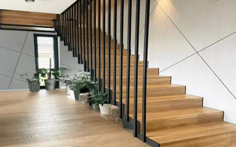 Nowoczesna klatka schodowa w domu z drewnianymi stopniami i czarną, metalową balustradą. Wokół rośliny w doniczkach.