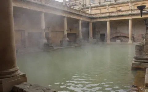 Dampf steigt aus dem Becken der römischen Therme auf. Säulen und Bögen umgeben das Wasser, in dem Enten schwimmen.