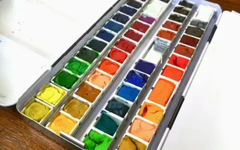 Paleta z różnymi rodzajami farb plastycznych: akwareli w kostkach, od żółci, przez zielenie, po brązy i błękity.