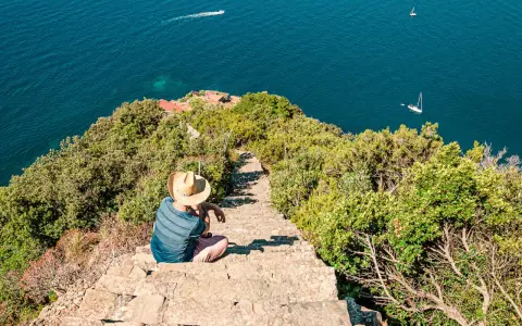 Un uomo con cappello di paglia seduto su una scalinata di pietra guarda il mare blu. Un sentiero scende verso il mare, suggerendo come arrivare a Monesteroli.