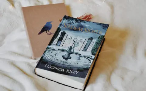 Okładka książki "Siedem Sióstr" Lucindy Riley na białym tle. Obok leży brązowy notes z wizerunkiem kruka.
