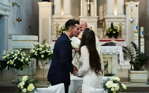 Para całuje się podczas ślubu konkordatowego w kościele, z księdzem w tle.
