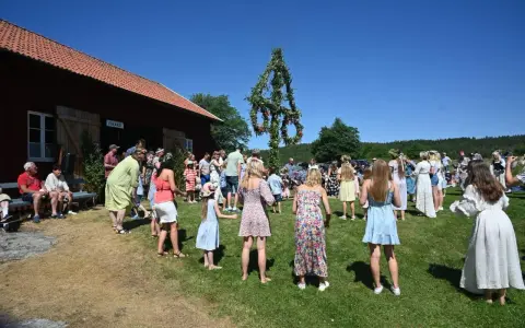 Folk dansar runt midsommarstången på midsommar afton. En vacker sommardag med klarblå himmel.