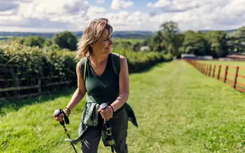Donna sorridente con bastoncini da nordic walking, che lavora i muscoli coinvolti in un paesaggio verdeggiante.