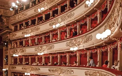 Widownia La Scali, pełna widzów w lożach, oczekuje na rozpoczęcie spektaklu. Bogaty repertuar teatru przyciąga miłośników sztuki.