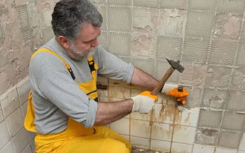 Un homme en salopette jaune utilise un marteau pour enlever le carrelage mural, révélant le plâtre en dessous.