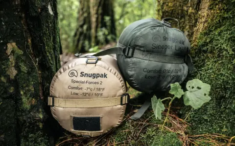 Dwa śpiwory Snugpak, jeden beżowy (komfort -7°C), drugi zielony (komfort 0°C), leżą na leśnym poszyciu.