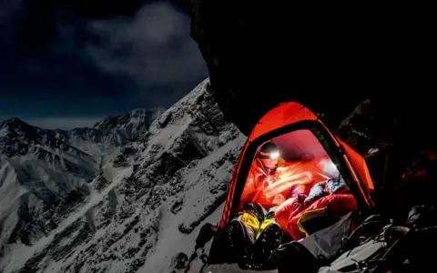 Nocny obóz na zboczu himalajskiego parbatu. W pomarańczowym namiocie rozświetlonym czołówką odpoczywa alpinista, otoczony surowym, ośnieżonym krajobrazem.