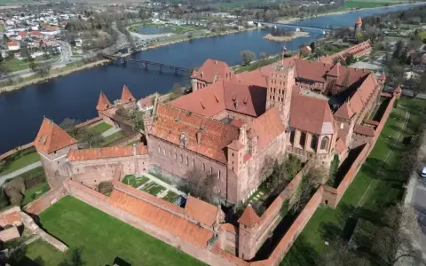Potężny zamek krzyżacki, największy zamek w Europie, wznosi się nad rzeką Nogat. Czerwona cegła i liczne wieże tworzą imponujący widok.