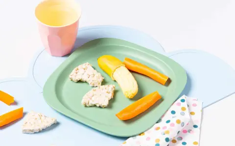Baby-gerechte Snacks: Reiswaffeln, Karottensticks und eine halbe Banane auf einem grünen Teller.
