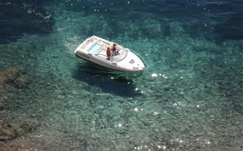 Deux personnes dans un bateau blanc, au moteur en panne, dérivent sur une eau turquoise et claire, révélant les fonds rocheux.