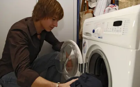 Junger Mann lädt Wäsche in die Waschmaschine. Bald werden die Sitzkissen frisch gewaschen sein.