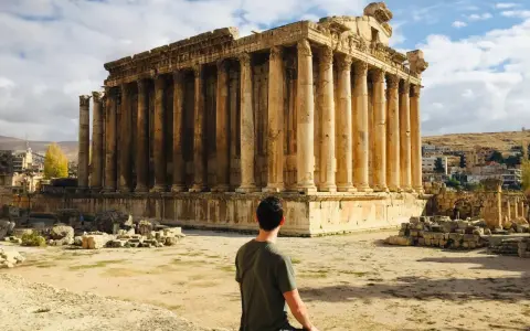 A man sits contemplating the grandeur of Roman ruins in Lebanon, bathed in sunlight under a cloudy sky.