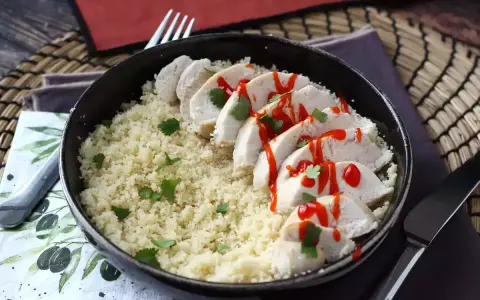 Delizioso petto di pollo in padella, affettato e servito su un letto di couscous, guarnito con prezzemolo e un filo di salsa piccante.