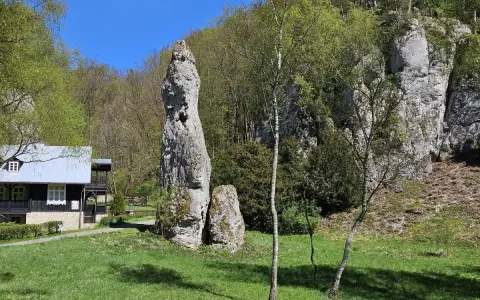 Imponująca skała Igła Deotymy góruje nad zieloną polaną, obok której stoi drewniany domek.